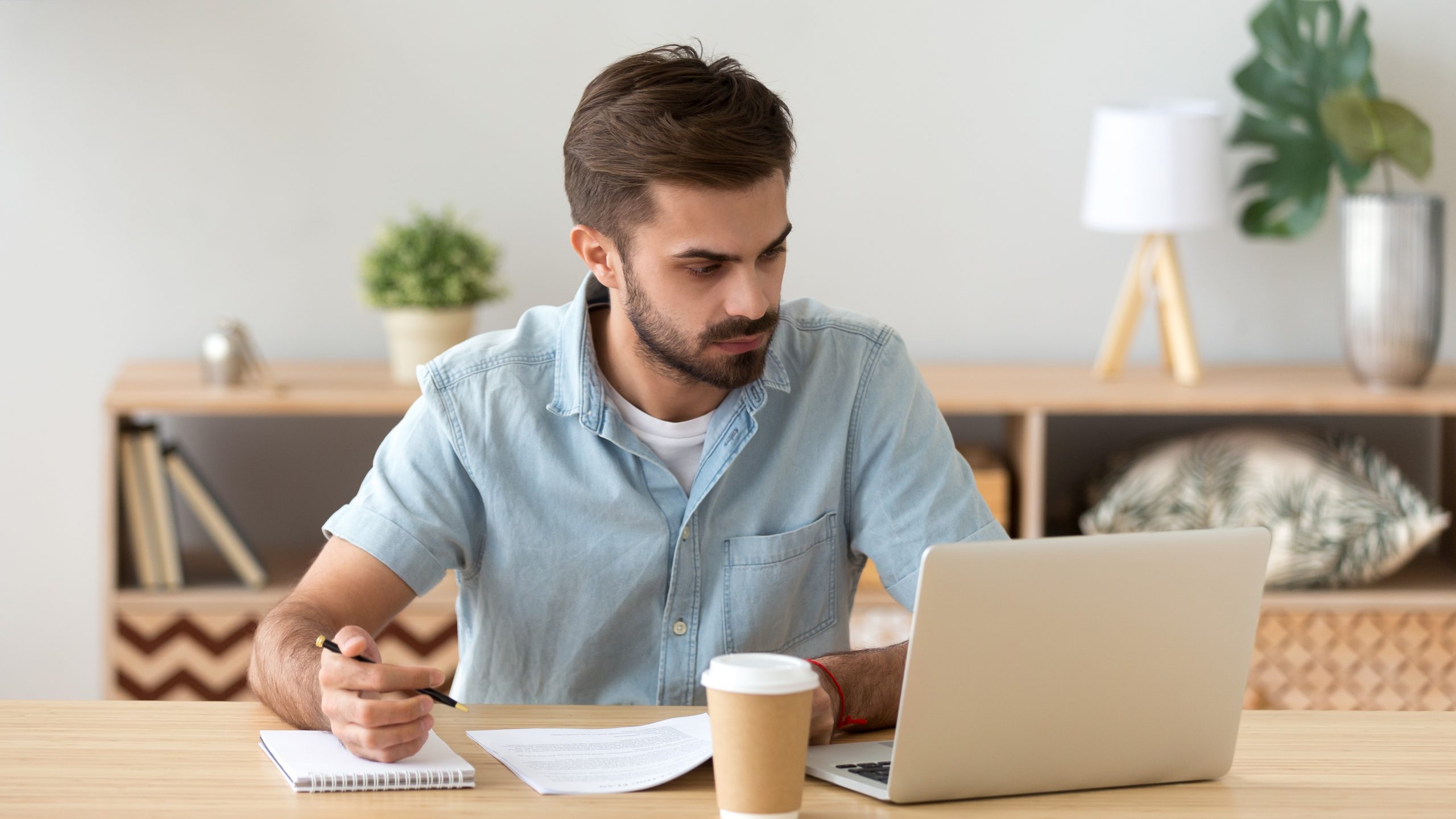 Serious young man looking at laptop making notes working with documents, doing paperwork, online research, focused male student studying in internet writing down information in notebook, e-learning
