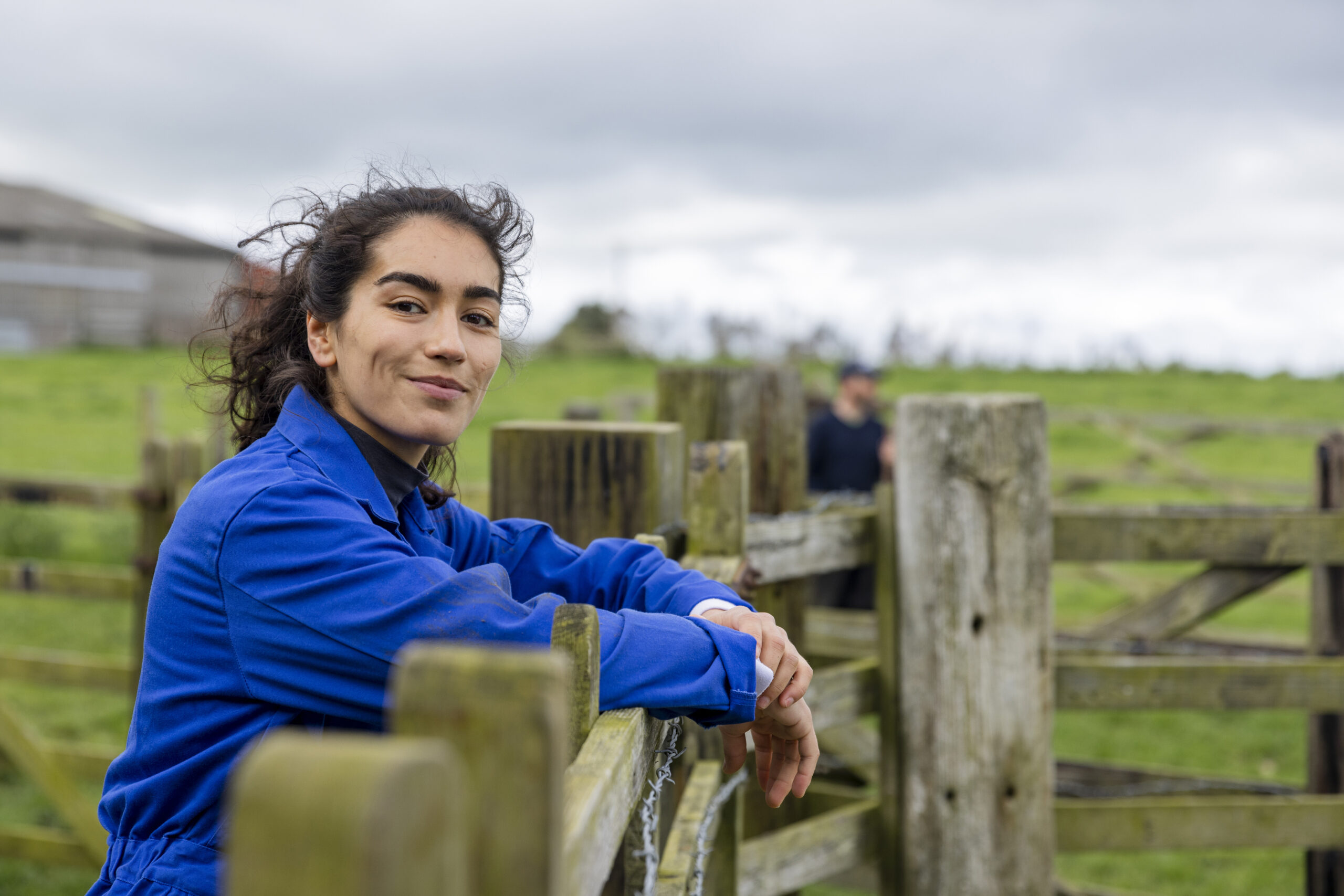 Confident in Her Line of Work A portrait of a young female farmer wearing overalls, standing and leaning against a fence at the farm she works at in Embleton, North East England while looking at the camera and smiling.